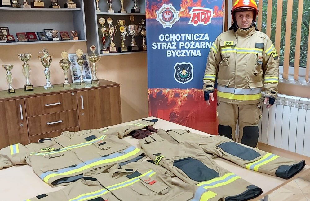 Ubrania strażackie ułożone na stole, po prawej strażak w umundurowaniu, w tle puchary