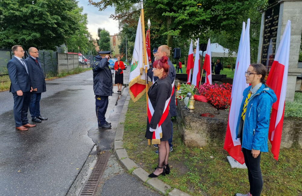 Kilka osób oddaje hołd przed pomnikiem, w tle flagi biało-czerwone