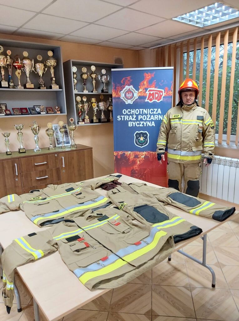 Ubrania strażackie ułożone na stole, po prawej strażak w umundurowaniu, w tle puchary