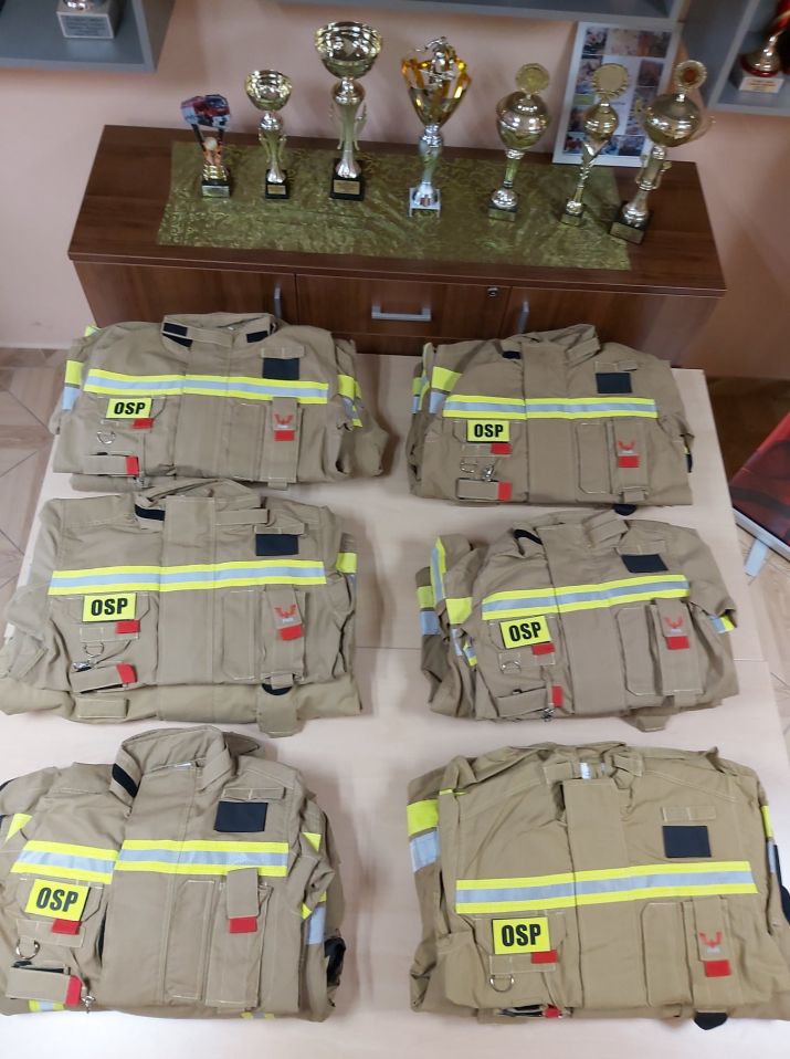 Sześć kompletów ubrań strażackich ułożonych na stole, w tle puchary