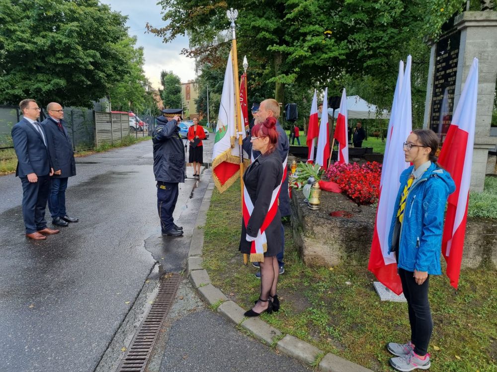 Kilka osób oddaje hołd przed pomnikiem, w tle flagi biało-czerwone