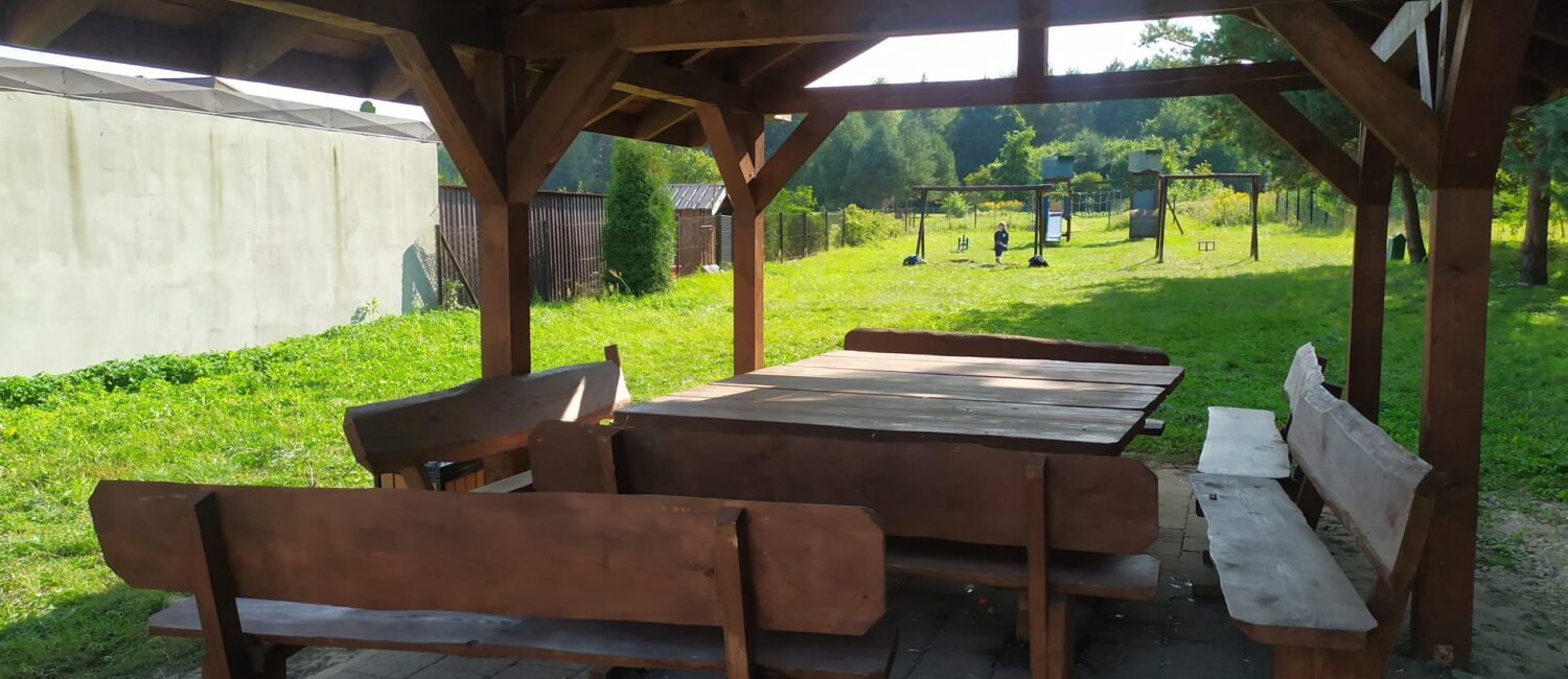 Drewniana altana, stół drewniany i ławki, w tle huśtawki dla dzieci