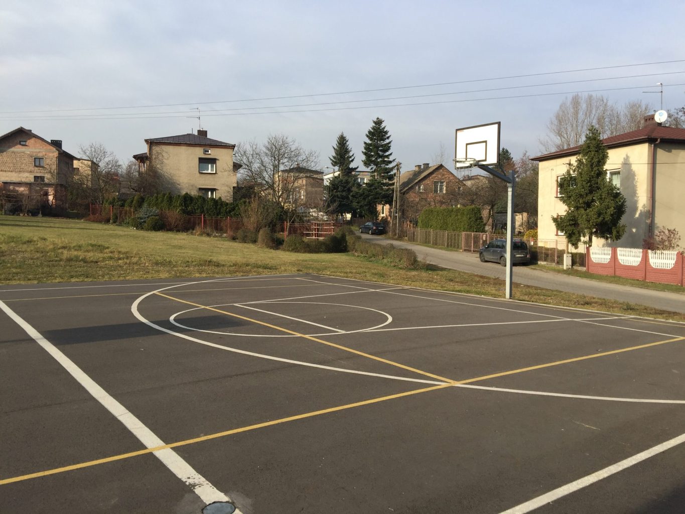 Boisko asfaltowe do gry w koszykówkę, w tle domy jednorodzinne