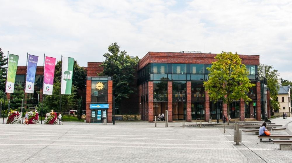 Budynek Miejskiej Biblioteki Publicznej w Jaworznie, przed nim od lewej cztery flagi na masztach, po prawej jawor