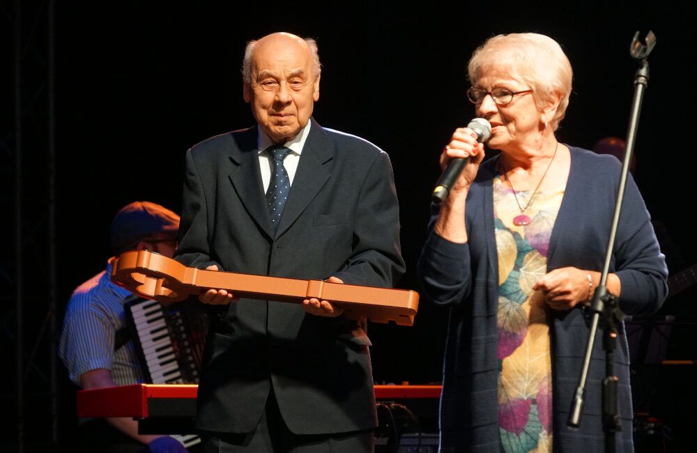 Starszy mężczyzna trzymający symboliczny klucz oraz starsza kobieta, która mówi do mikrofonu