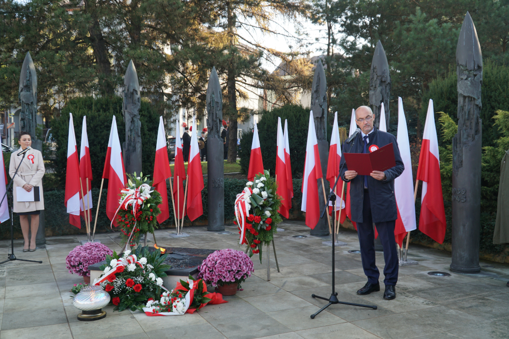 Mężczyzna przemawia do mikrofonu, w rękach trzyma czerwoną teczkę, w tle biało-czerwone flagi oraz kwiaty