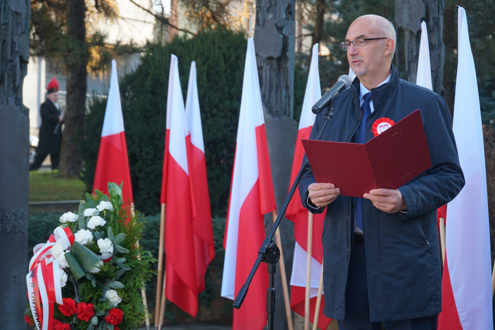 Mężczyzna przemawia do mikrofonu, w rękach trzyma czerwoną teczkę, w tle biało-czerwone flagi