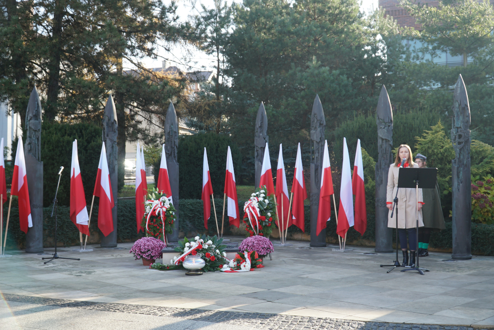 Kobieta przemawiająca do mikrofonu pod Pomnikiem Niepodległości. W tle biało-czerwone kwiaty i flagi