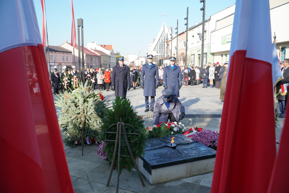 Czterech mundurowych składa kwiaty pod pomnikiem niepodległości, na pierwszym planie planie dwie biało-czerwone flagi