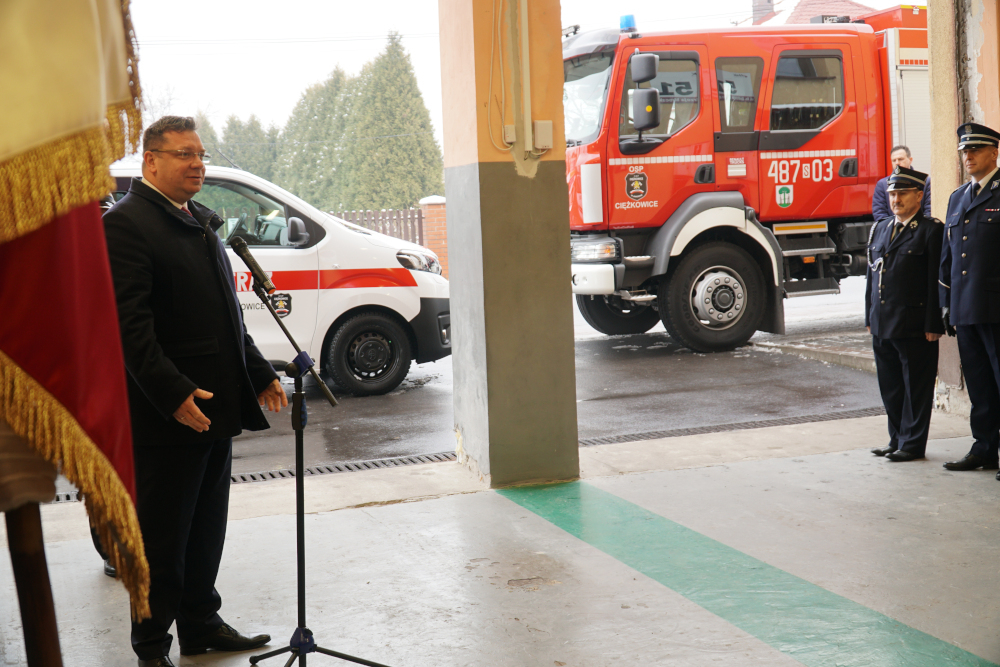Mężczyzna przemawiający do mikrofonu, w tle wozy strażackie