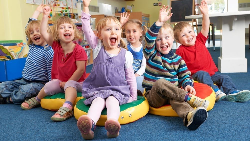 Grupa dzieci siedząca na dywanie w sali i podnosząca rączki do góry