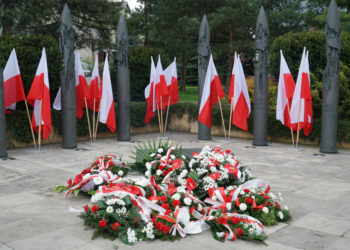 Wiązanki biało-czerwonych kwiatów ułożone pod pomnikiem, w tle flagi biało-czerwone