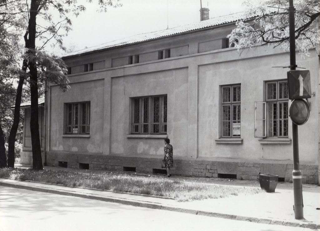 Czarno-białe zdjęcie budynku, przed budynkiem spaceruje kobieta