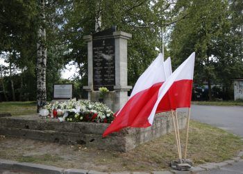 Pomnik przyozdobiony kwiatami, po prawej trzy flagi biało-czerwone