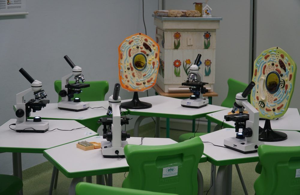 Klasa szkolna. Na pierwszym planie stoły z mikroskopami i zielone krzesła