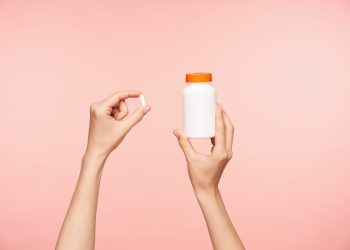 Przycięte zdjęcie uniesionych zadbanych dłoni kobiety trzymającej białą pigułkę i butelkę z pomarańczową pokrywką, przyjmujących witaminy, pozując na różowym tle