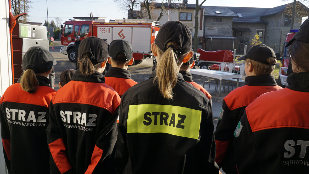 Grupa dzieci stojąca tyłem, ubrana w kurtki z napisem STRAŻ, na tle wozu strażackiego