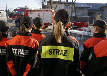 Grupa dzieci stojąca tyłem, ubrana w kurtki z napisem STRAŻ, na tle wozu strażackiego