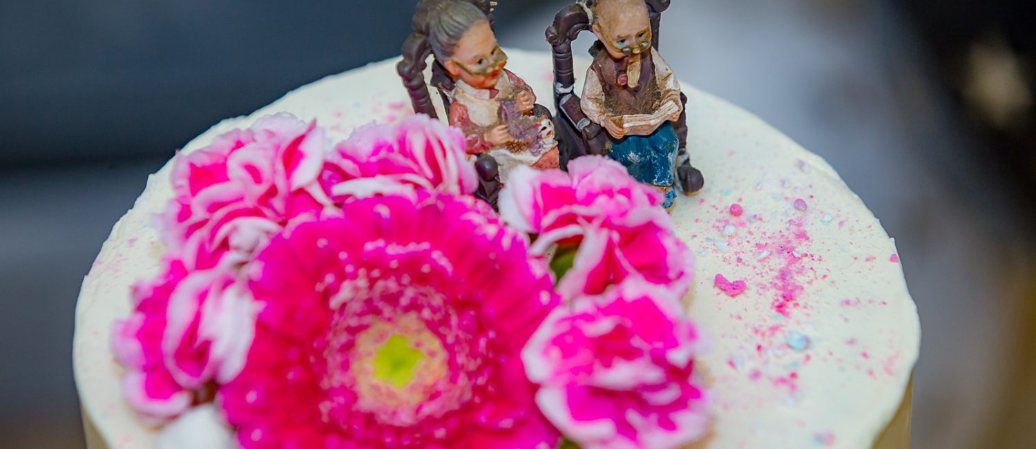 Biały tort z różowymi kwiatami oraz figurkami babci i dziadka.