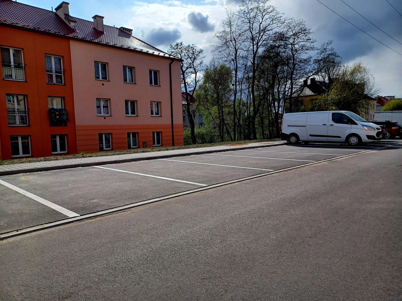 Fragment drogi z wyznaczonymi miejscami parkingowymi, w tle bloki mieszkalne