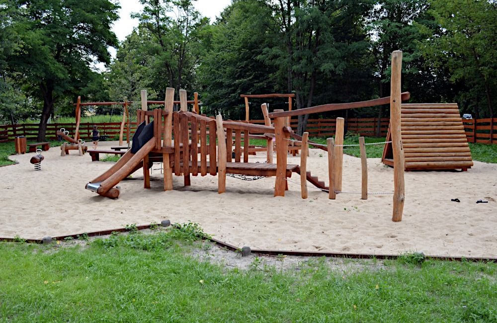 Plac zabaw wykonany z naturalnych elementów drewnianych na podłożu piasek w tle zielone drzewa