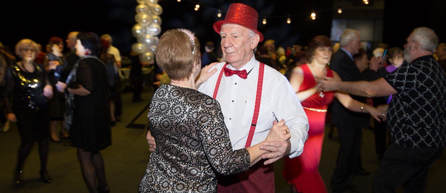 Mężczyzna w czerwonej czapce, czerwonej muszce, i czerwonych szelkach tańczy na sali z kobietą, która obrócona jest tyłem do obiektywu.