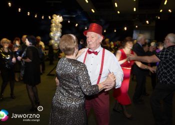 Mężczyzna w czerwonej czapce, czerwonej muszce, i czerwonych szelkach tańczy na sali z kobietą, która obrócona jest tyłem do obiektywu.